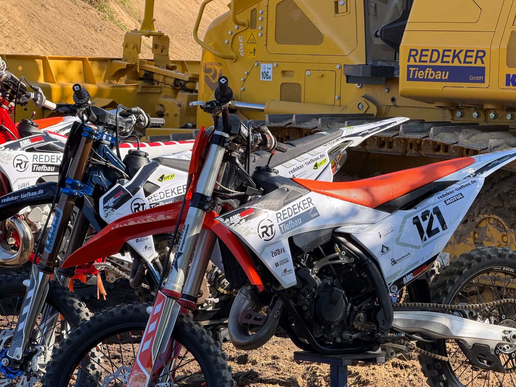 Motocross-Motorrad im Redeker-Tiefbau-Design vor einem Baufahrzeug der Redeker Tiefbau GmbH