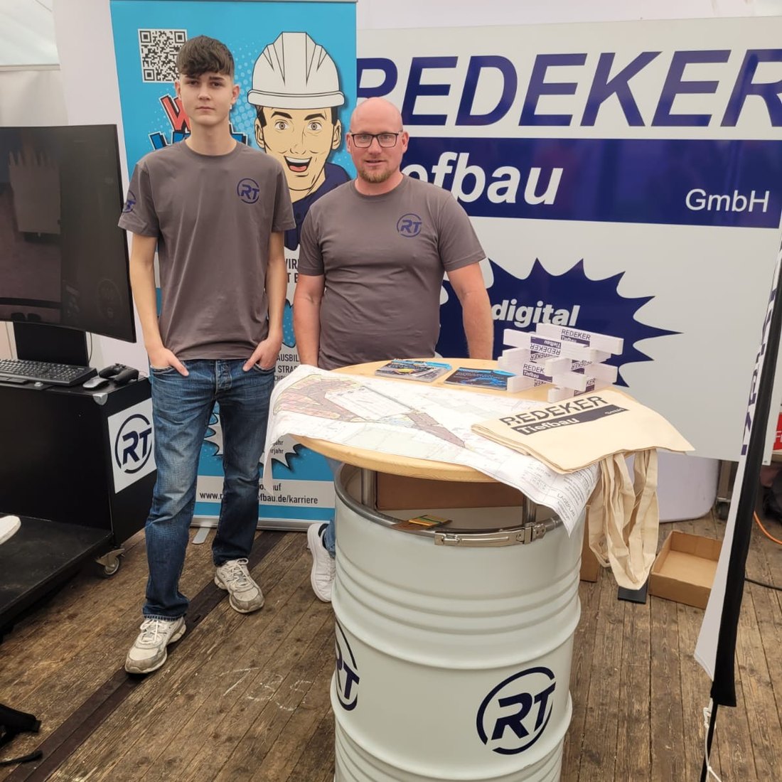 Ausbilder und Auszubildender der Redeker Tiefbau GmbH am Messestand vor dem Firmen-Roll-up