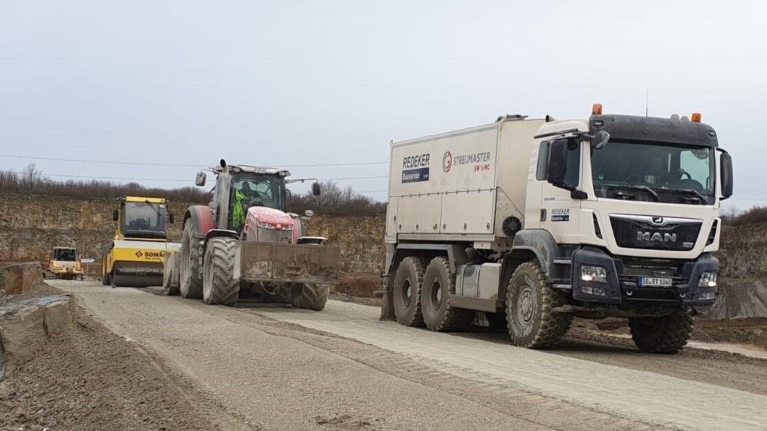 Redeker-Tiefbau-Streumaster-Lkw und Traktor mit Walze bei der Bodenstabilisierung im Steinbruch