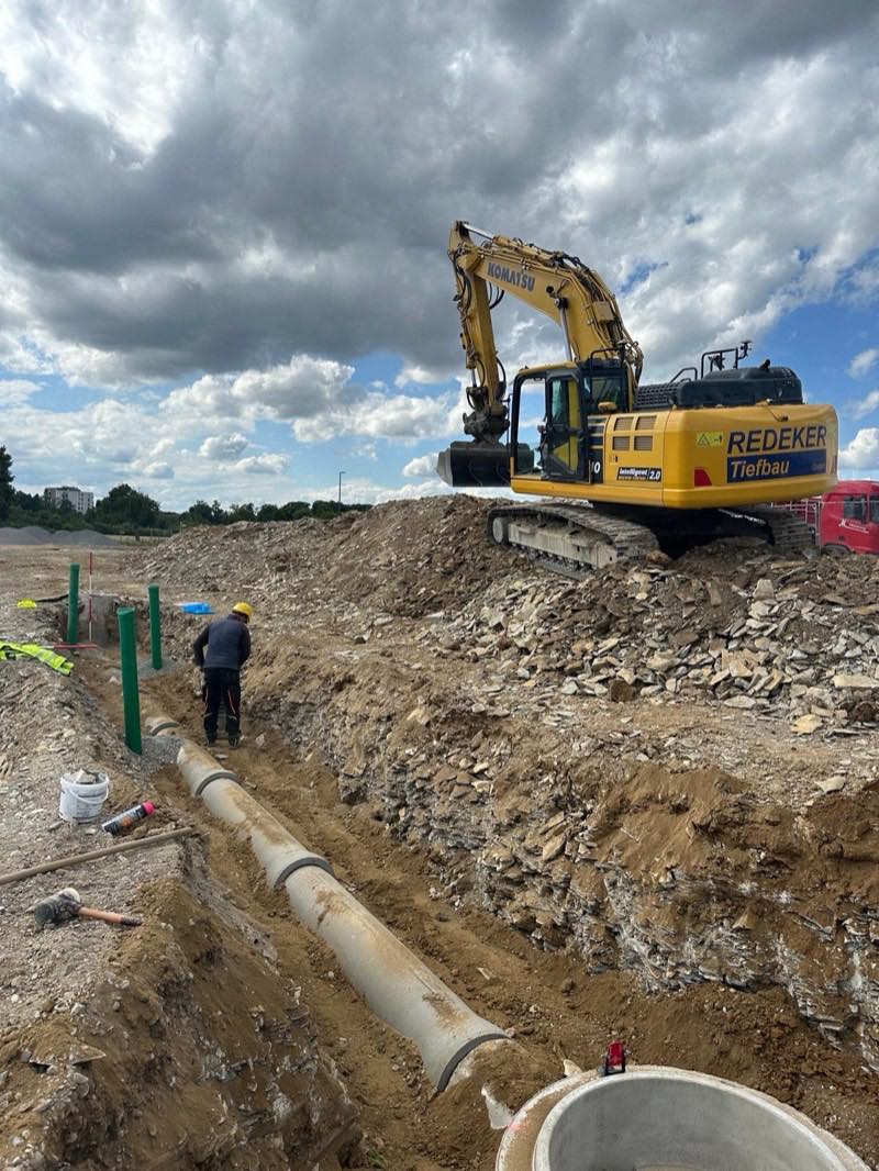 Kanalbauer verlegt ein Betonrohr im Rohrgraben, dahinter ein Redeker-Tiefbau-Kettenbagger